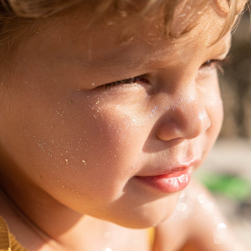 close up of child showing glitter suncreen on face and cheeks with gold bio glitter 50+ sunscreen for kids